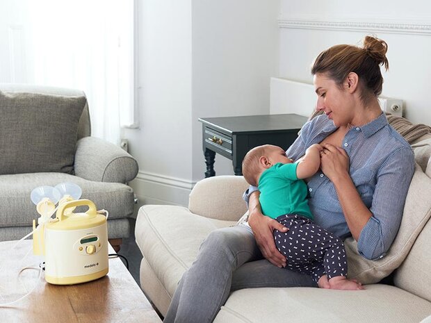 Mutter sitzt auf dem Sofa und stillt ihr Baby im Wohnzimmer, daneben steht eine Milchpumpe auf dem Tisch